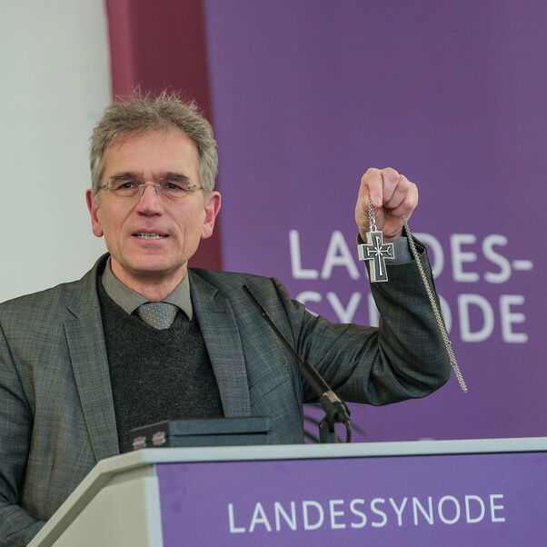 Eine männlich lesbare Person an einem Rednerpult mit einem Kreuz in der Hand