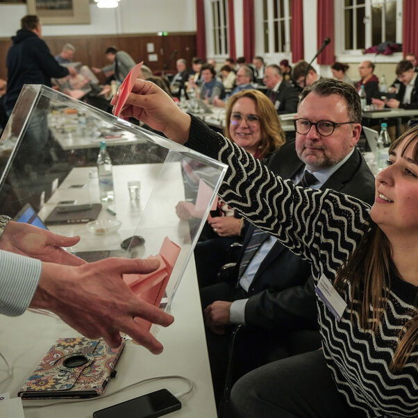 Eine weiblich lesbare Person wirft einen Stimmzettel in eine Wahlurne