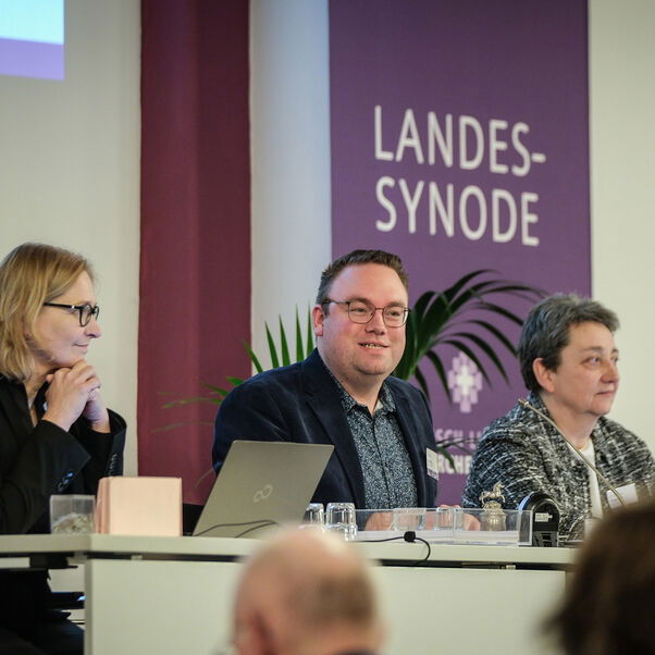 Drei Personen an einem Tisch in einem Sitzungssaal