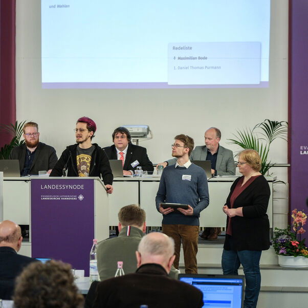 Mehrere Personen an Tischen und stehend in einem Sitzungssaal