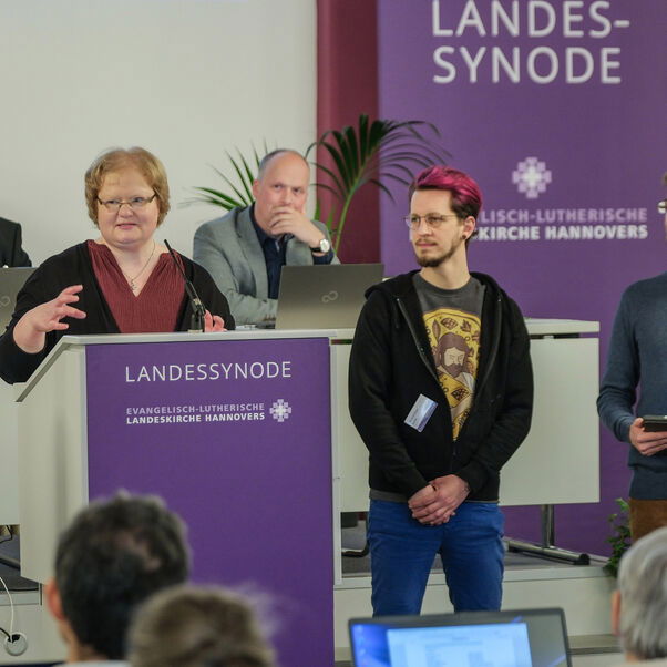 Mehrere Personen an Tischen und stehend in einem Sitzungssaal