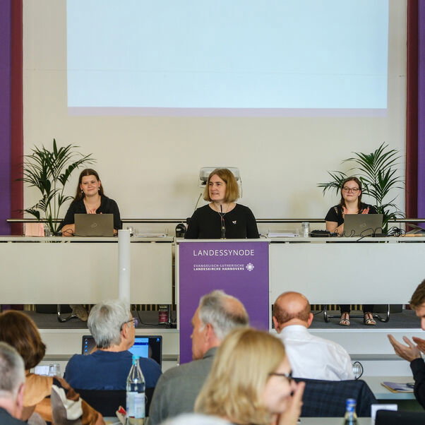 Drei Personen sitzen an einem Tisch in einem Konferenzsaal