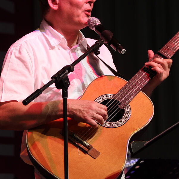 Eine als Mann lesbare Person mit Gitarre singt in ein Mikrofon