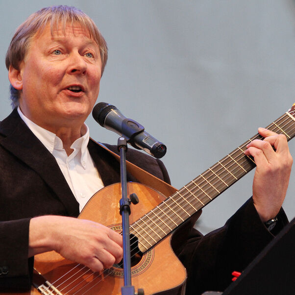 Eine als Mann lesbare Person mit Gitarre singt in ein Mikrofon