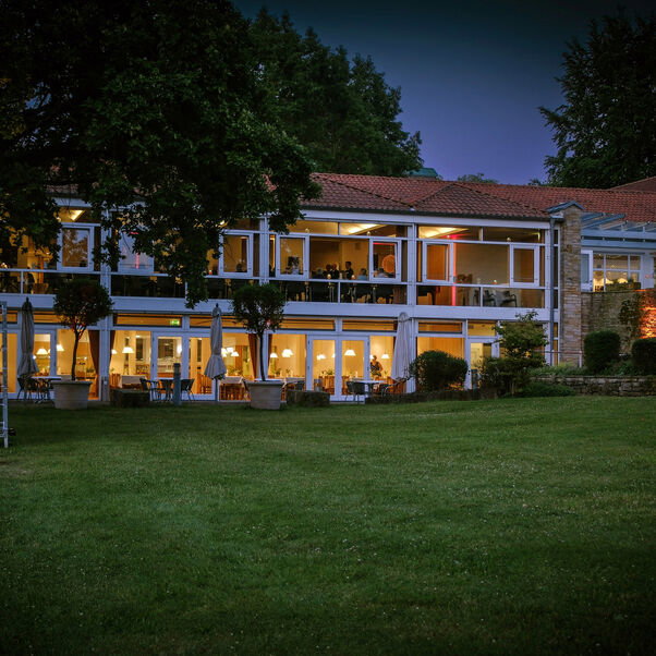 Es ist die Evangelische Akademie Loccum in dunkeln zu sehen. Im Vordergrund ist eine große Grünfläche. Im Hintergrund die Fensterfronten von dem großen Speisesaal im Erdgeschoss sowie der Galerie im ersten Stock.