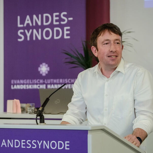 Eine männlich gelesene Person in weißem Hemd an einem Redepult. 
