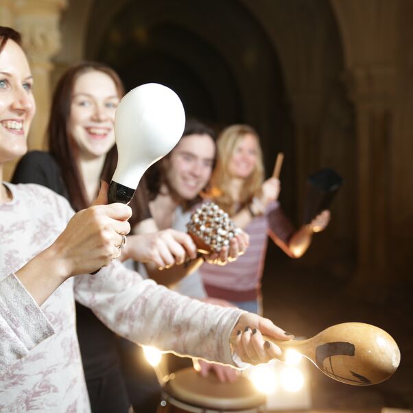 Das Bild zeigt mehrere weiblich lesbare Personen, die mit großen Rasseln Musik machen. 