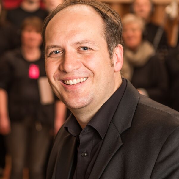 Das Bild zeigt den Oberkörper eines männlich lesbaren, lächelnden Menschen mit kurzen schwarzen Haaren und schwarzem Oberhemd. Im Hintergrund steht eine Gruppe von Menschen in Choraufstellung.