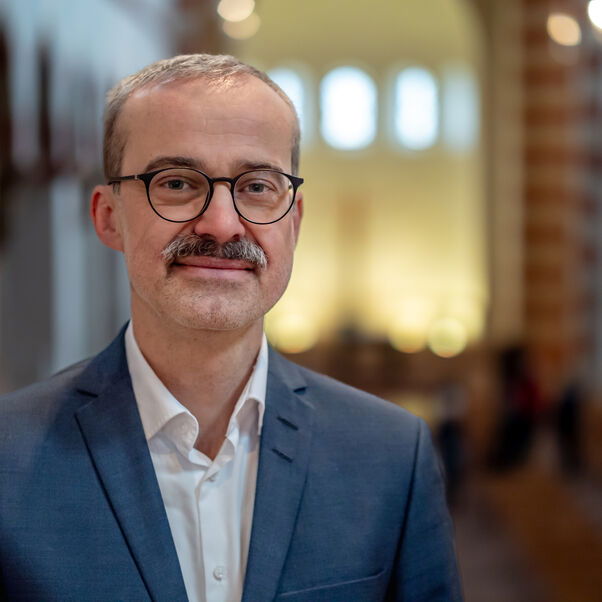 Das Bild zeigt eine männlich lesbare Person mit blauem Sakko, weißem Hemd, kurzen dunklen Haaren, Schnurrbart und Brille. 