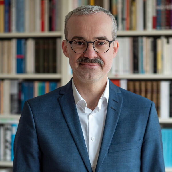 Das Bild zeigt eine männlich lesbare Person mit blauem Sakko, weißem Hemd, kurzen dunklen Haaren, Schnurrbart und Brille. 