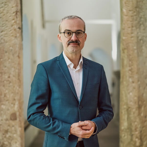 Das Bild zeigt eine männlich lesbare Person mit blauem Sakko, weißem Hemd, kurzen dunklen Haaren, Schnurrbart und Brille. 