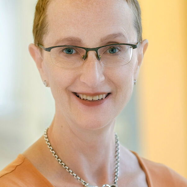 Das Bild zeigt eine weiblich lesbare Person mit sehr kurzen rotbraunen Haaren, einer Brille mit Oberfassung, einem orangefarbenen Oberteil und einer silbernen Kette.