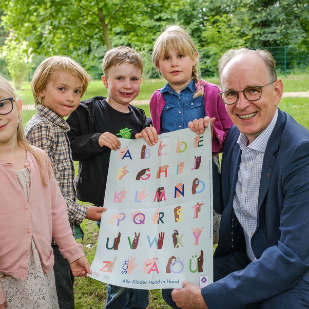 Landesbischof Ralf Meister und vier Kinder zeigen das Poster mit dem Fingeralphabet.