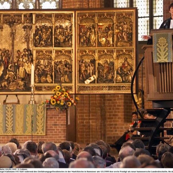 Eine weiblich zu lesende Person steht im Talar auf einer Kanzel und spricht zu einer vollbesetzten Kirche.