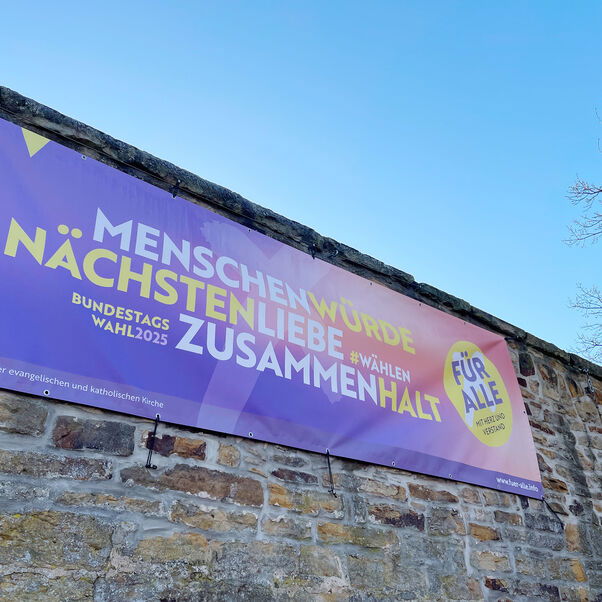 An einer hohen Steinmauer hängt ein Banner, auf dem die Worte Menschenwürde, Nächstenliebe, Zusammenhalt stehen.