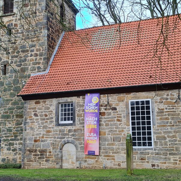 An einer kleinen, steinernen Kirche hängt ein rosa-lila Banner, auf dem die Worte Menschenwürde, Nächstenliebe, Zusammenhalt stehen.