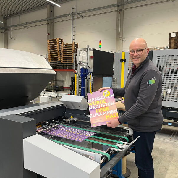An einem großen Industriedrucker steht eine Person, die ein Plakat in Händen hält, auf dem die Worte "Menschenwürde-Nächstenliebe-Zusammenhalt" stehen.