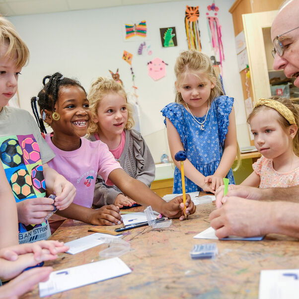 Eine männlich zu lesende Person knete mit Kindern, die an einem Tisch sitzen, Figuren.