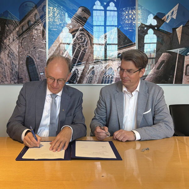 Zwei männlich zu lesende Personen sitzen an einem Tisch und eine Person unterschreibt ein Dokument, das auf dem Tisch liegt.