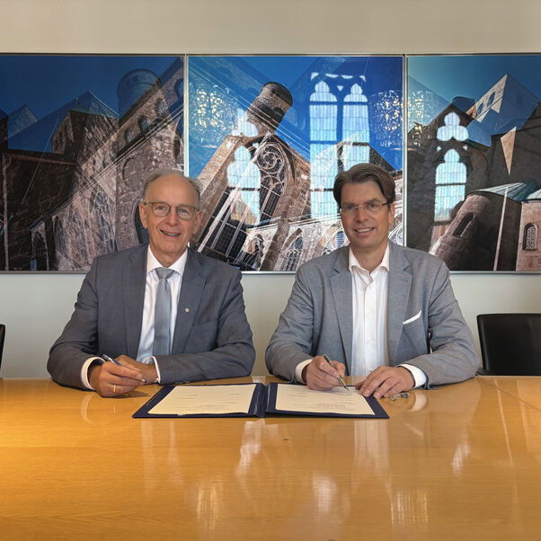 Zwei männlich zu lesende Personen sitzen an einem Tisch und vor ihnen liegt ein Dokument zur Unterschrift.