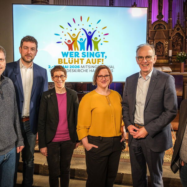 Eine Gruppe von als Männer und Frauen lesbaren Personen steht in einer Kirche vor einer Leinwand mit dem Logo zum Mitsingfestival „Wer singt, blüht auf!“