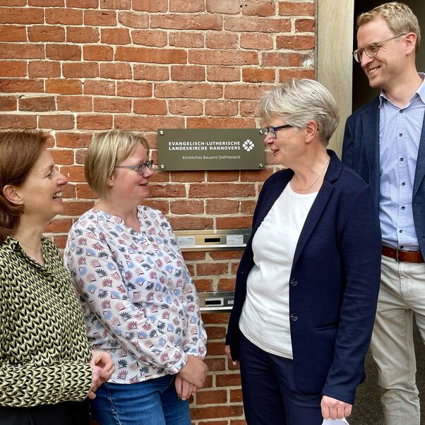 Vier Personen stehen vor einer Eingangstür, in ihrer Mitte das frisch enthüllte neue Schild mit folgender Schrift: Evangelisch-lutherische Landeskirche Hannovers Kirchliches Bauamt Ostfriesland.
