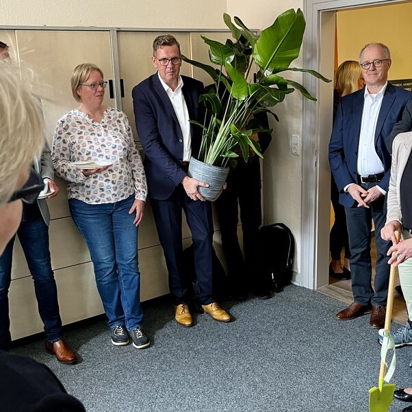 Mehrere Personen stehen in einem Büro, eine Person hält eine große Topfpflanze in Händen, eine andere eine kleine Spielzeug-Schaufel als Geschenk.