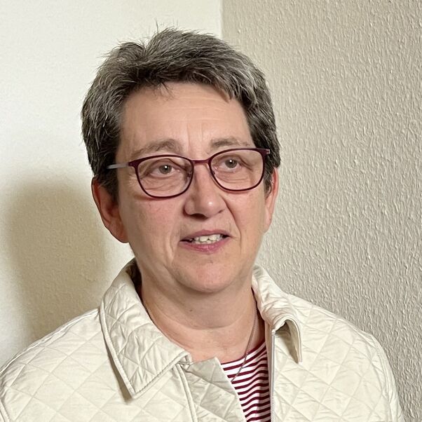 Weiblich lesbare Person mit kurzen grauen Haaren, Brille und heller Jacke.