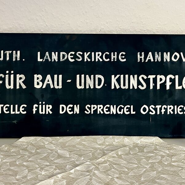 Ein abgeschraubtes Türschild, schwarz mit weißer Schrift: "Ev.-luth. Landeskirche Hannovers Amt für Bau- und Kunstpflege Außenstelle für den Sprengel Ostfriesland