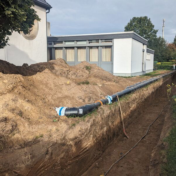 Neben einem ausgehobenen Graben vor einer Kirche liegen schwarze Fernwärmerohre.