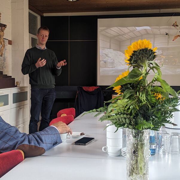 Ein Mann steht vor einer Projektionswand und hält einen Vortrag während ein anderer Mann davor sitzt und ihm zuhört.