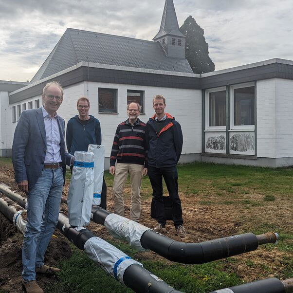 Vier Menschen stehen vor einem Gebäude und schauen sich sich große schwarze Rohre an.