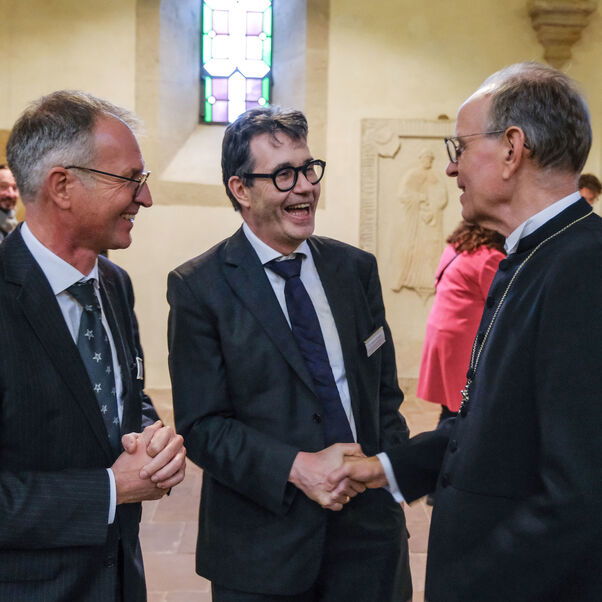 Drei männlich zu lesende Personen begrüßen sich in einer Kirche.