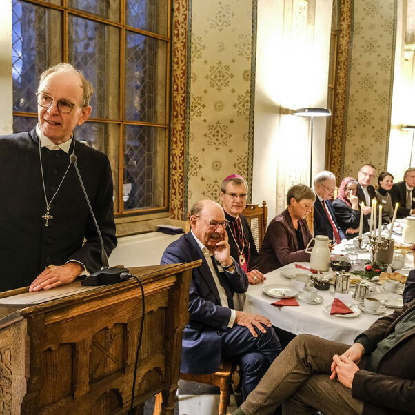 Eine männlich zu lesende Person steht an einem Pult und spricht zu eine Gruppe von Personen, die an einem gedeckten Tisch sitzen.