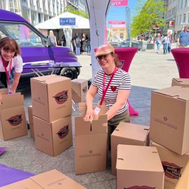 Zwei weiblich zu lesende Personen bauen Papphocker in einer Fußgängerzone auf.