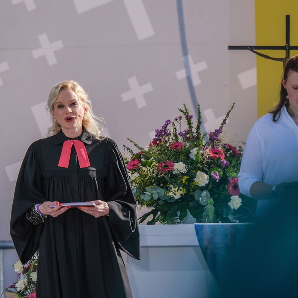 Zwei weiblich zu lesende Personen stehen vor einem Altar. Eine Person trägt einen Talar.