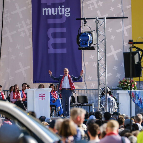 Eine männlich zu lesende Person steht auf einer Bühne und hebt zum Segen beide Arme.
