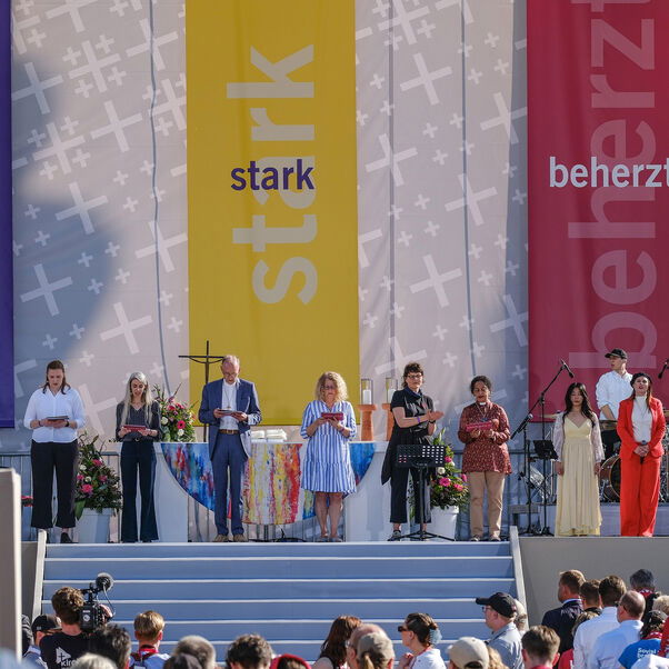 Auf einer Bühne stehen Personen nebeneinander. Im Hintergrund sind drei farbige Banner zu sehen.