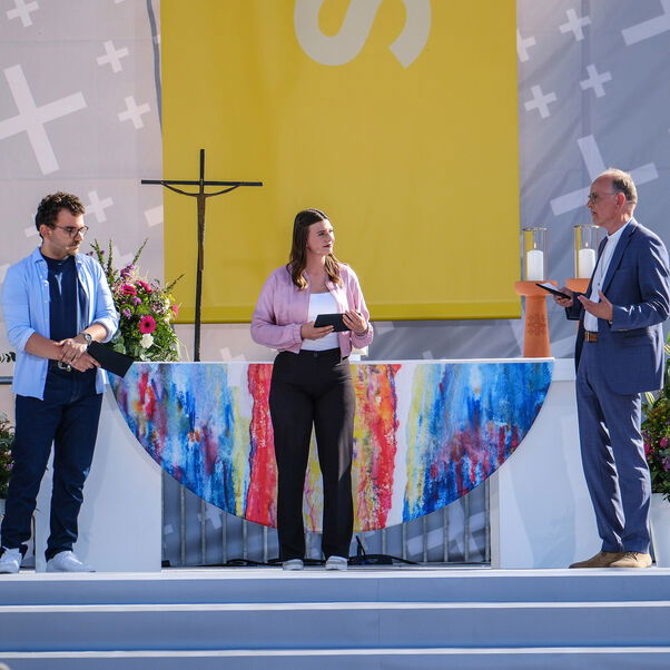 Zwei männlich und eine weiblich zu lesende Person stehen vor einem Altar und unterhalten sich während sie in Mikrofone sprechen.