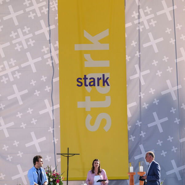 Zwei männlich und eine weiblich zu lesende Personen stehen vor einem Altar und unterhalten sich.