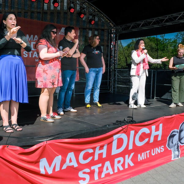 Eine Gruppe von Personen steht auf einer Bühne. Eine weiblich zu lesende Person spricht in ein Mikrofon.