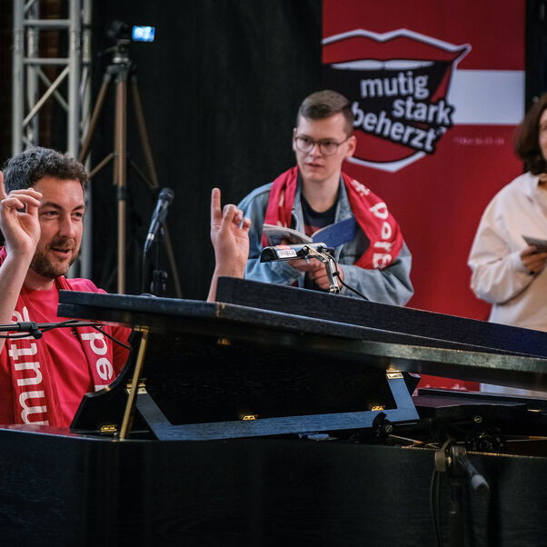 Eine männlich zu lesende Person sitzt an einem Flügel und dirigiert eine Gruppe von Personen, die um den Flügel herumstehen.