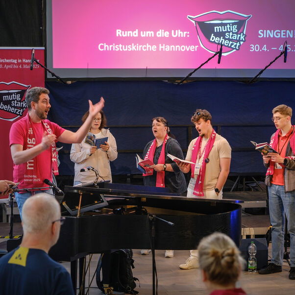 Eine männlich zu lesende Person steht an einem Flügel und dirigiert eine Gruppe von Personen, die um den Flügel herumstehen.