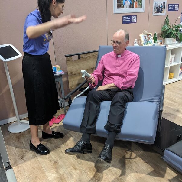 Eine männlich zu lesende Person sitzt auf einem elektrisch verstellbaren Stuhl. Eine weiblich zu lesende Person erklärt ihm die Funktionsweise.