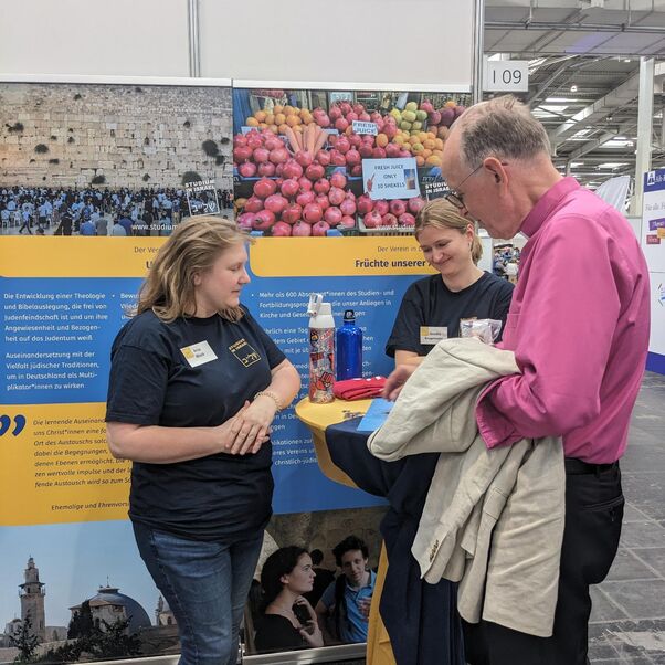 Eine männlich zu lesende Person steht an einem Messestand und unterhält sich mit einer weiblich zu lesenden Person.