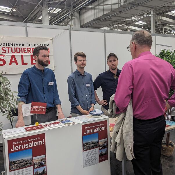 Eine männlich zu lesende Person steht am Tresen eines Messestandes und unterhält sich mit drei männlich zu lesende Personen, die hinter dem Tresen stehen.