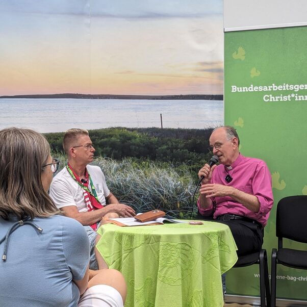 Zwei männlich zu lesende Personen sitzen an einem kleinen Tisch und eine Person spricht in ein Mikrofon.
