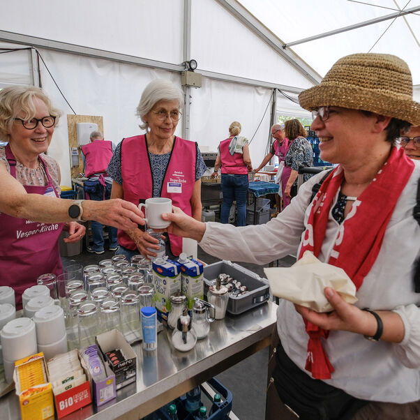 Weiblich zu lesende Personen geben an einem Tresen Getränke an andere Personen aus, die in einer Schlange stehen.