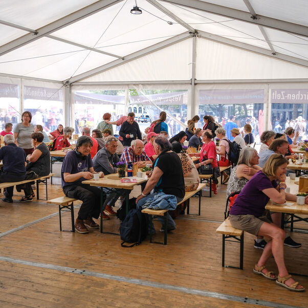 Weiblich und männlich zu lesende Personen sitzen an Bierzeltgarnituren in einem Zelt.