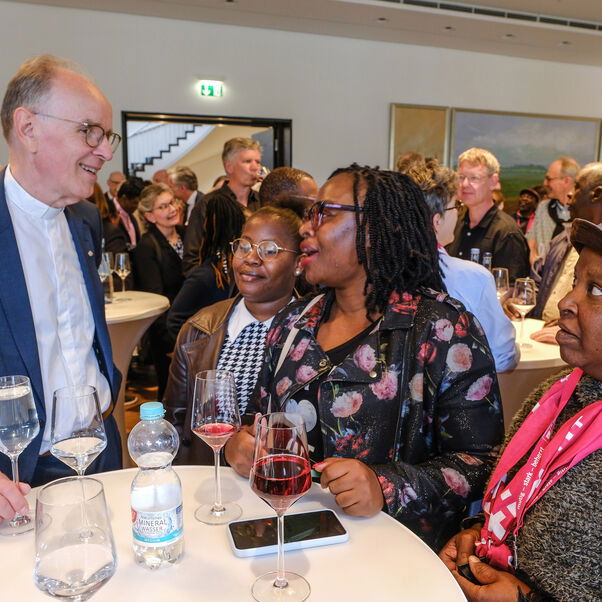 Zwei weiblich zu lesende Personen unterhalten sich an einem Stehtisch mit einer männlich zu lesenden Person.